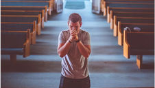 A man in gray shirt praying in an empty church, kneeling between pews. Soft light fills the room, creating a serene atmosphere.