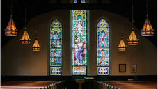 Stained glass windows in a church with biblical scenes. Warm lighting from hanging lamps creates a serene, reverent atmosphere over wooden pews.