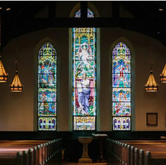 Stained glass windows in a church with biblical scenes. Warm lighting from hanging lamps creates a serene, reverent atmosphere over wooden pews.