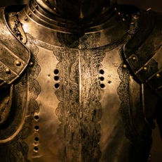 Close-up of ornate, dark metallic medieval armor with intricate engravings. The setting is dimly lit, highlighting the detailed craftsmanship.