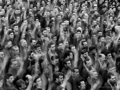 A black-and-white image shows a large crowd of men in motion, many raising their hands, creating a dynamic, blurred effect. Mood appears solemn.
