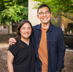 Two people smiling outdoors, standing closely with one arm around the other. They're in casual outfits, surrounded by greenery.