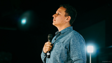 Man in blue shirt holds a microphone, looking thoughtful on a dimly lit stage. Bright light in background. Mood is contemplative.