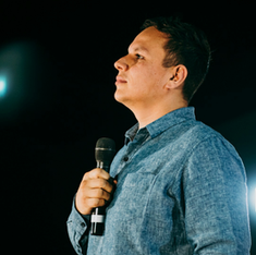 Man in blue shirt holds a microphone, looking thoughtful on a dimly lit stage. Bright light in background. Mood is contemplative.