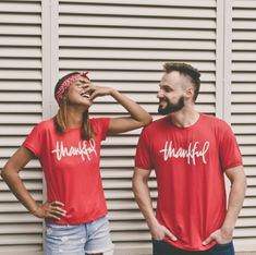 Two people in red "thankful" shirts smile and laugh. Background features white shutters. One wears a red bandana and denim shorts. Mood is joyful.
