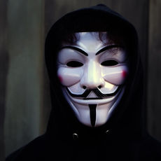 A person in a black hoodie wears a white Guy Fawkes mask, standing against a dark, blurred background, creating a mysterious mood.