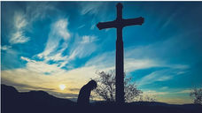 Silhouette of a person bowing by a large cross at sunset. Blue sky with scattered clouds sets a tranquil, reflective mood.