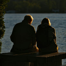 Two people sit on a dock, silhouetted against a lake at sunset. The scene is peaceful, with soft golden light and trees in the background.