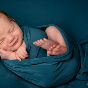 Newborn baby smiling, wrapped in a teal blanket against a teal background, conveying a peaceful and joyful mood.