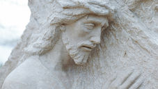 Stone sculpture of a sorrowful man with beard and crown of thorns, leaning against a cross. The background is a cloudy sky.