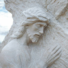 Stone sculpture of a sorrowful man with beard and crown of thorns, leaning against a cross. The background is a cloudy sky.