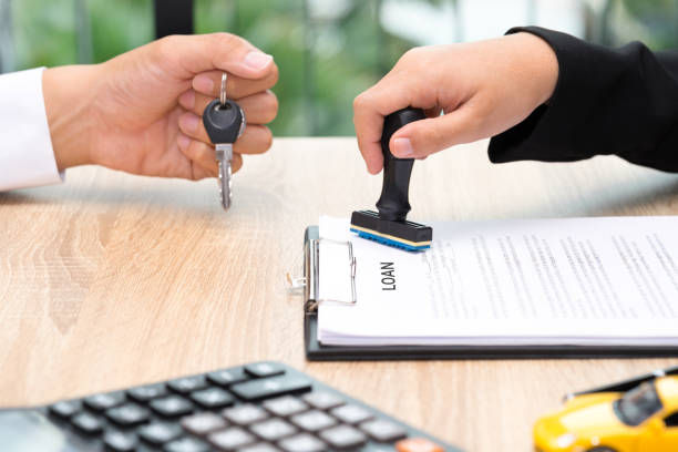 Close-up of a person stamping a loan document on a clipboard while another hand holds car keys, symbolizing an approved car loan. A calculator and a small toy car are visible on the table.