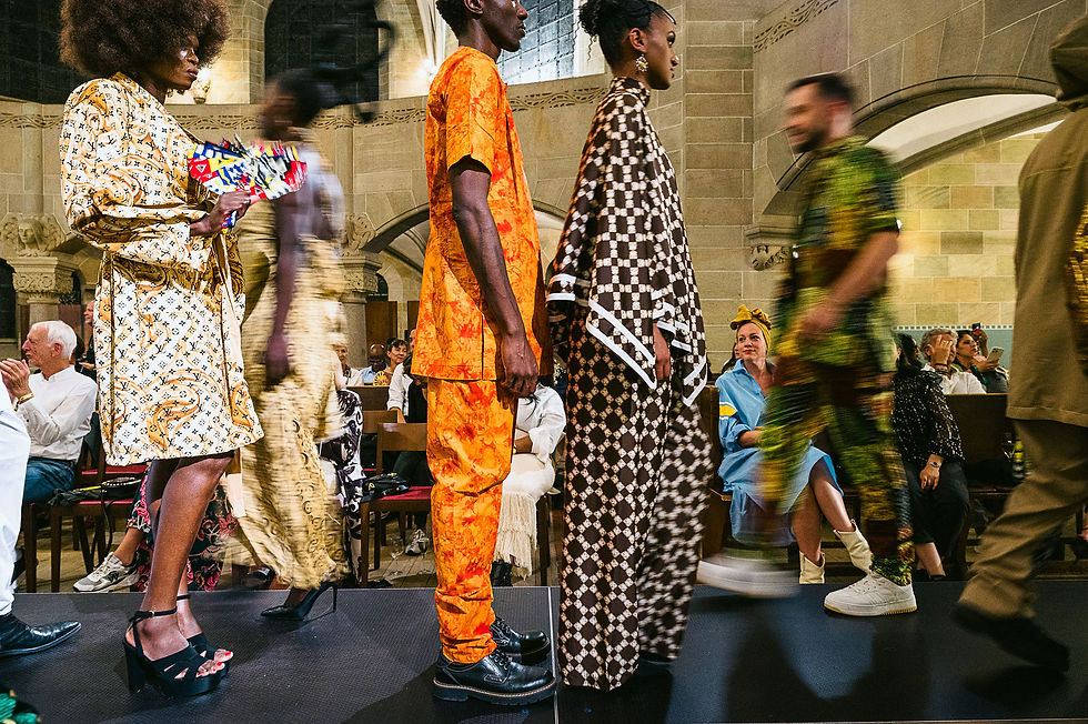 Models in bright patterned outfits walk a runway in a stone hall as blurred guests watch, creating a lively fashion-show scene.
