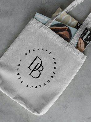 White tote bag with "Deanna Becraft Photography" logo, containing magazines with visible covers. Set on a gray concrete surface.