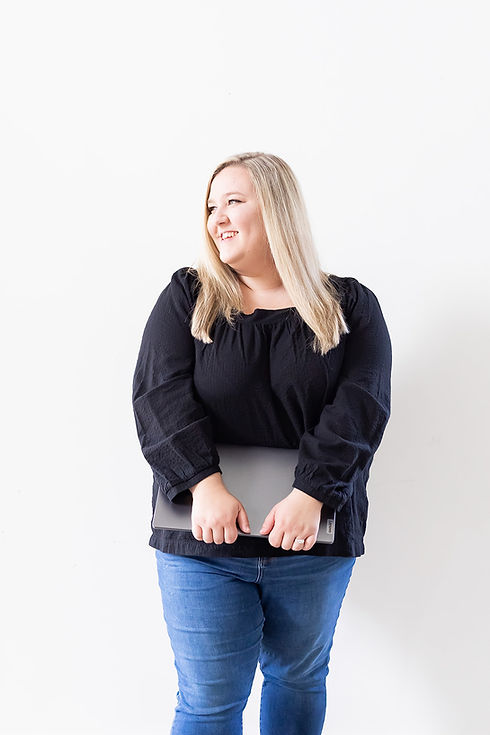 female brand and web designer in a black long sleeve shirt and jeans, holding laptop in front of her, looking off to the side smiling in front of a white wall