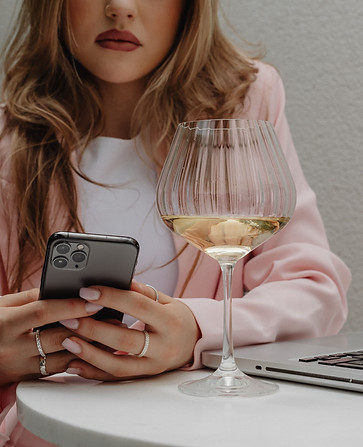 woman in a pink blazer at a table on her phone, drinking wine