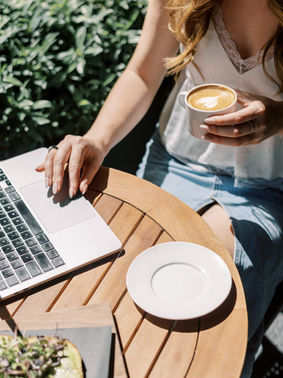 Woman in a white top and denim skirt using a laptop and holding a latte at a wooden table outdoors. Sunny, with greenery in the background.