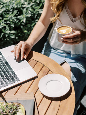 Woman in a white top and denim skirt using a laptop and holding a latte at a wooden table outdoors. Sunny, with greenery in the background.