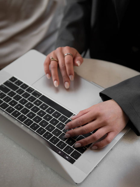 Hands with rings typing on a laptop keyboard. The person wears a dark sleeve. The setting is minimal, with a light-colored surface.