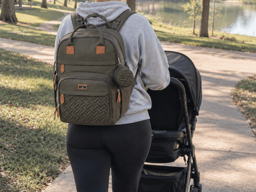 Woman in gray hoodie and black leggings pushes a stroller on a sunny park path beside a lake, wearing a large olive backpack.