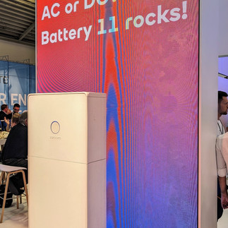 Das Bild zeigt ein modernes Batteriespeichersystem, das auf InterSolar 2024 München ausgestellt ist. Dahinter ist ein großer Bildschirm mit dem Text „AC oder DC? Battery 11 rockt!“ zu sehen. Die Kulisse scheint ein belebter Messestand mit vielen Menschen zu sein. Das Gerät hat ein schlichtes, minimalistisches Design.
