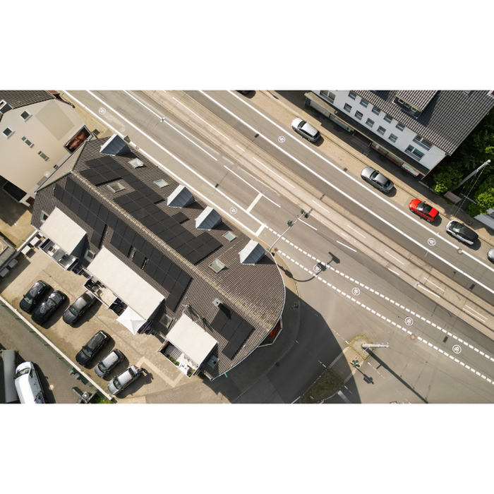 "Luftaufnahme eines Gebäudes mit dunklem, geometrischem Dach, ausgestattet mit einer von Algatec Solar gebauten Solaranlage. Die Anlage bietet 29 kWp mit 69 Modulen à 420 Wp und zeigt die Expertise von Algatec Solar in nachhaltiger Energie. Autos parken auf einer Seite, eine gepflegte grüne Umgebung auf der anderen unterstreicht Effizienz und Umweltfreundlichkeit.