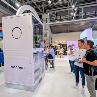 Eine Gruppe von Menschen steht während der InterSolar 2024 München um einen hohen weißen Stand mit der Aufschrift „Sonnen“. Die Innenausstattung des Standes, in der das Speichersystem von Sonnen ausgestellt ist, ist durch transparente Paneele sichtbar. Im Hintergrund ist ein moderner, gut beleuchteter Veranstaltungsraum mit zusätzlichen Sitzgelegenheiten und Ausstellungen zu sehen.