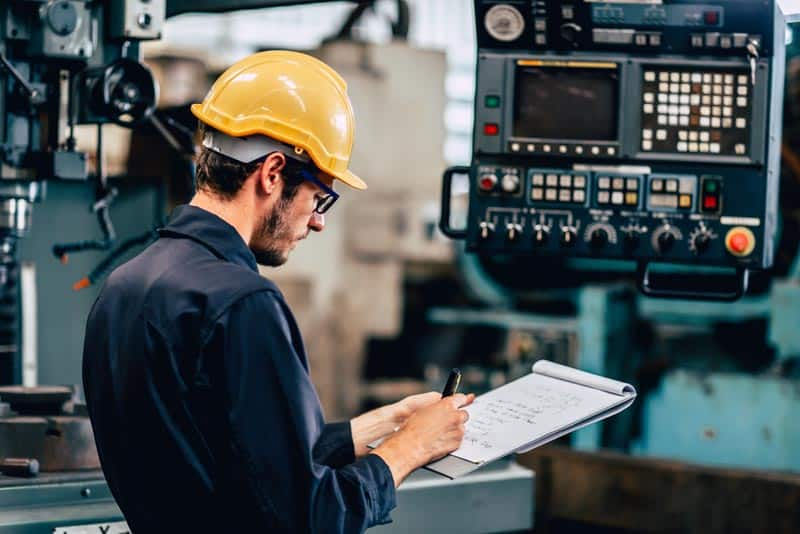 Engineer in yellow helmet inspecting machinery in a factory, writing notes