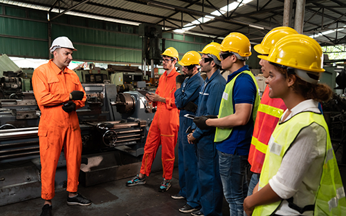 Engineer in orange jumpsuit giving instructions to group of workers at factory.