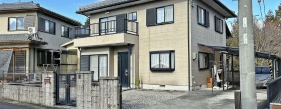 The photo shows a two-story house with a beige exterior and a black roof. The house has a balcony on the second floor and a small front yard with a gate and a driveway. The sky is blue with some white clouds. The house appears to be well-maintained and in a quiet residential neighborhood.
