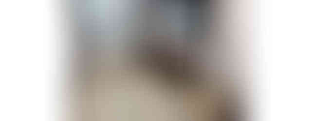 A photo of a Japanese-style living room with tatami mats and sliding doors. The room features a low table and a television. There is a door leading to another room in the background. The ceiling has a ceiling light and a clock.