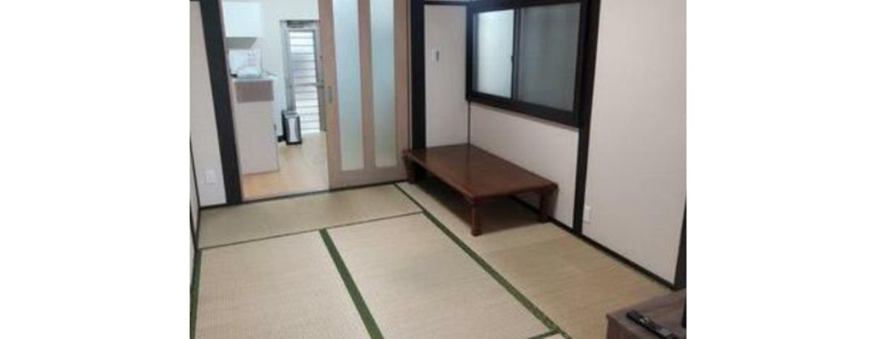 A photo of a Japanese-style living room with tatami mats and sliding doors. The room features a low table and a television. There is a door leading to another room in the background. The ceiling has a ceiling light and a clock.