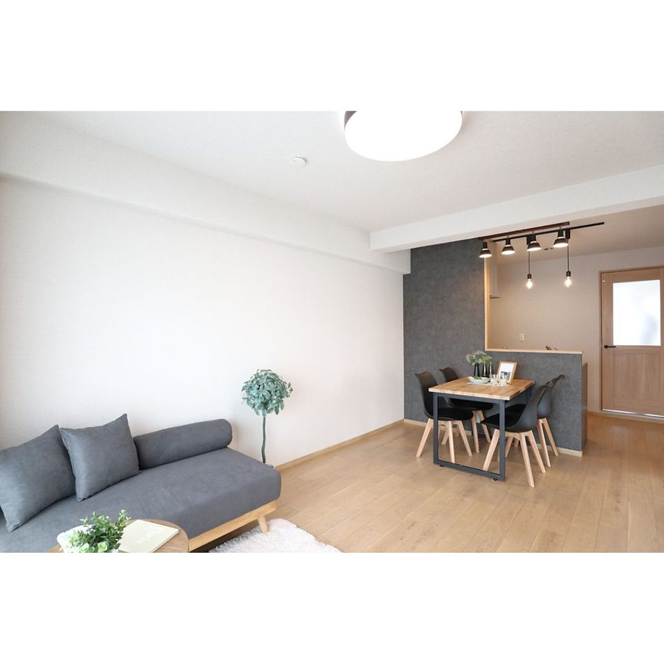  A bright and airy living room with a minimalist aesthetic. A gray sofa with pillows sits opposite a dining table with four black chairs. The floor is a light wood and the walls are white. There is a door to the right of the dining table and a small balcony with a plant to the left.
