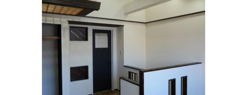 An interior view of a modern Japanese house entrance. The space features a dark wood-paneled ceiling with exposed beams and recessed lighting. A white wall with a contrasting dark border frames a black door with a small window. A half-wall with decorative cutouts separates the entrance area from another room. The floor is covered in light brown wood-look flooring.