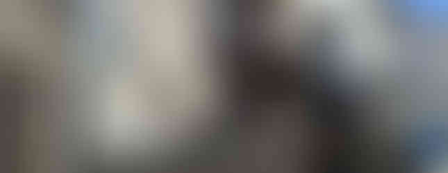 The image shows a two-story apartment building with a white facade and a red brick accent wall. The building has several balconies and a small courtyard in front. There are several windows on each floor, suggesting multiple units within the building. The building is located in a residential area with other houses visible in the background.