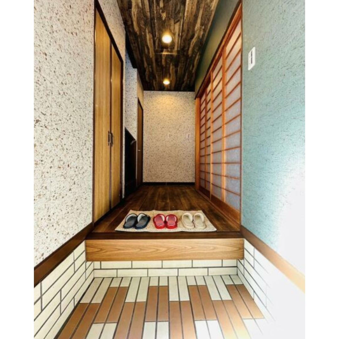 The image shows a traditional Japanese entranceway or genkan. There is a tiled floor with a pair of shoes on it and a small wooden bench. To the right, there is a built-in wooden shelf and cabinet. A sliding glass door leads to a small outdoor garden.