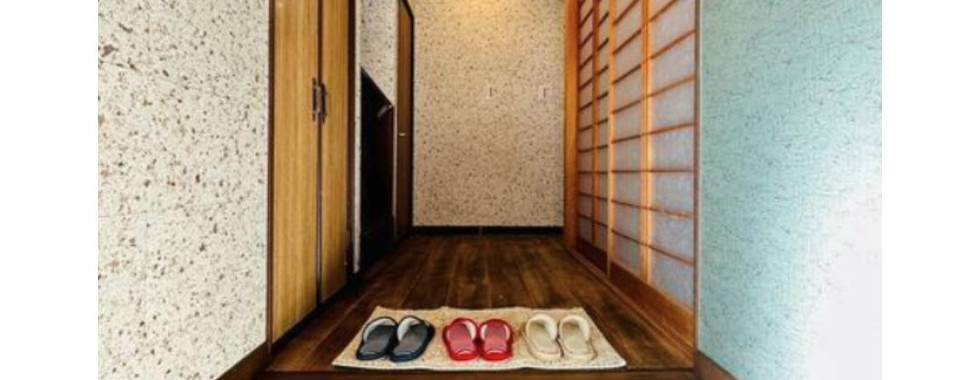 The image shows a traditional Japanese entranceway or genkan. There is a tiled floor with a pair of shoes on it and a small wooden bench. To the right, there is a built-in wooden shelf and cabinet. A sliding glass door leads to a small outdoor garden.