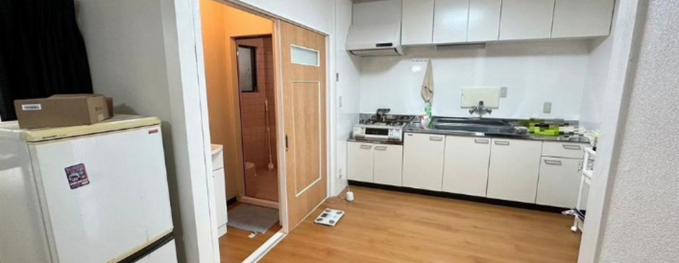 A compact kitchen and dining room area. Light brown wood flooring covers the space. On the left, a white refrigerator stands next to a sliding door, partially open to reveal a bathroom with a glimpse of a bathtub. On the right, a kitchen counter with white cabinets and a stainless steel sink occupies the wall.