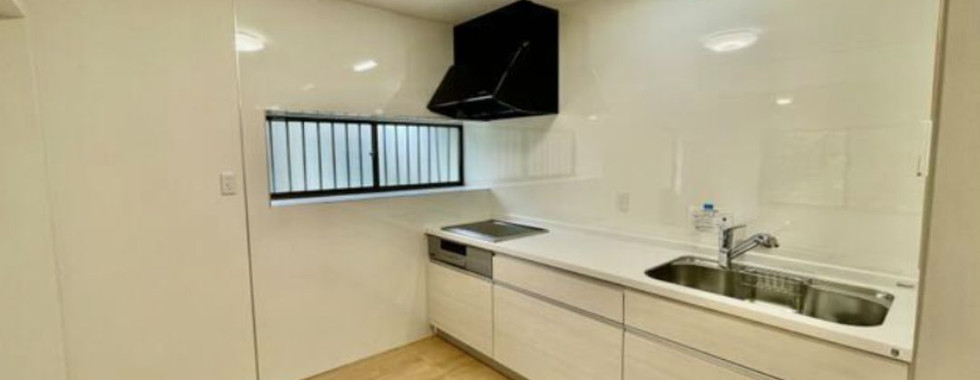 This photo shows a modern kitchen with white cabinets and a white countertop. There is a stainless steel sink with a faucet, a gas stovetop, and a range hood above it. The walls are tiled in white, and there is a window on the left side of the image.