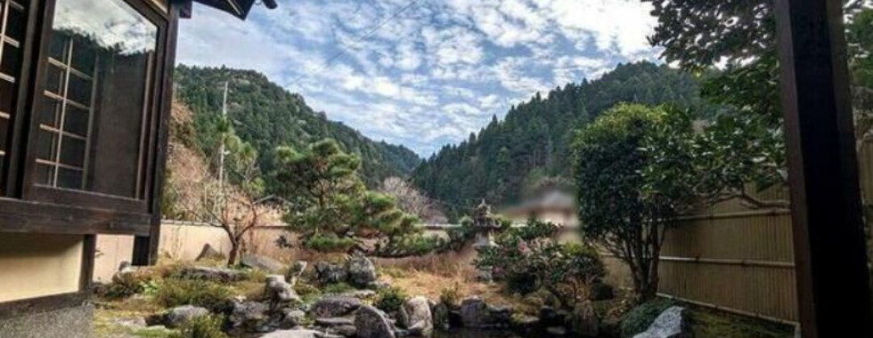 The photo shows a picturesque view of a Japanese garden from inside a traditional house. A small pond with rocks and a wooden bridge is visible in the foreground, surrounded by lush greenery. In the background, rolling hills covered in trees create a serene and peaceful landscape. The sky is a beautiful blue with fluffy white clouds.