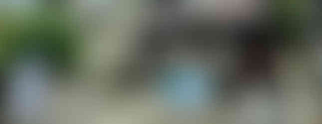    This photo shows a two-story house with a brown exterior. The house is located on a hill with a driveway leading up to the entrance. There is a small garden in front of the house and a wooden fence surrounding the property. The sky is cloudy.