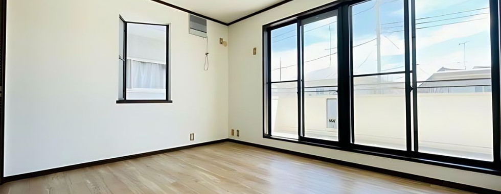 A bright and spacious room with light brown wooden flooring and white walls. Large windows with black frames offer ample natural light and a view to the outside. A small window with a curtain adds a touch of softness to the space. The room is unfurnished, highlighting its potential for various uses.