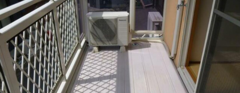 The photograph shows a balcony with a gray tiled floor and white metal railings. An air conditioning unit is mounted on the wall to the left. The balcony overlooks other buildings and houses in the neighborhood.