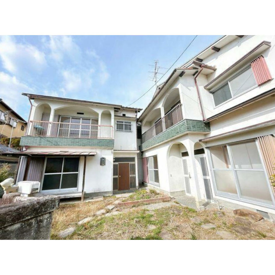 The photo shows a two-story house with a white facade and a balcony on the second floor. The house has a small front yard with a paved walkway and some landscaping.