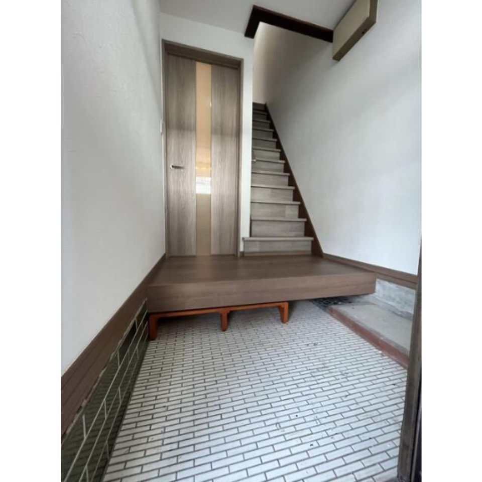 A Japanese-style entrance hall with a wooden floor and staircase. The hall has a modern sliding door on the left and a small wooden bench on the right. There is a pattern on the floor.