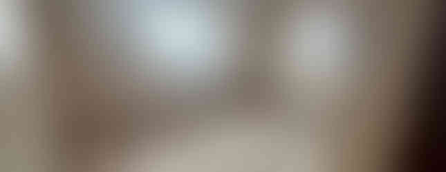 A carpeted bedroom with white walls and a ceiling fan. There are two windows with white curtains, a door, and a radiator in the room.