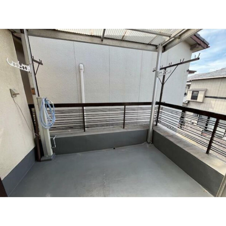 The photo shows a spacious balcony with a concrete floor. There is a metal railing and a covered roof overhead. A clothesline is attached to the wall, suggesting it's a functional outdoor space for laundry and other activities. The balcony looks clean and well-maintained.