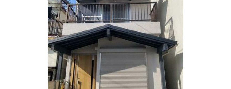 A photo of a modern, two-story house with a gray facade and a black roof. The house features a balcony on the second floor and a covered entranceway with a gray roller shutter door. The surrounding area includes a paved driveway, a small garden, and a utility pole with electrical wires.