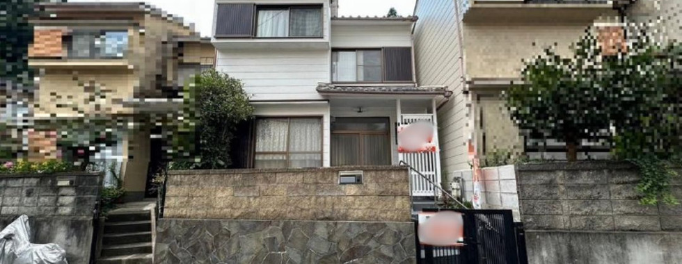 A two-story white house with a brown tiled roof. It's located on a street with other houses on either side. A low stone wall borders the front yard, and a black metal gate provides access to the entrance.
