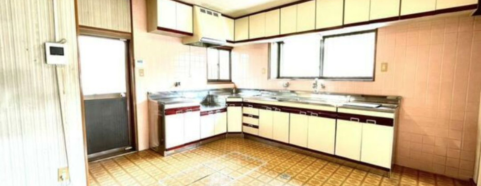 The photo shows a kitchen with white cabinets and a pink tiled wall. There is a sink with a window above it and a stovetop. The floor is covered in a brown and white checkered pattern. There is a door on the left side of the image leading to another room.
